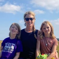 A white woman in a black sleeveless top and sunglasses with blond hair in a braid sitting between her two twin daughters, all three looking at the camera and smiling with blue sky and clouds in the background