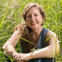 A middle aged white woman with long light brown curly hair pulled over her shoulder, wearing a blue and white pattered sleeveless top, sitting a field of tall green grass, smiling and looking at the camera