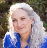 A white woman with long curly gray hair, wearing a blue top, looking at the camera and smiling