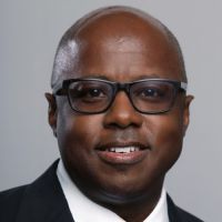 A bald, brown skin man wearing black glasses and a suit looking at the camera and smiling