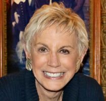A white woman with short gray hair in a pixie cut, wearing a black sweater and gold earrings, sitting in front of a blue painting, looking at the camera and smiling.