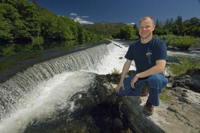 white man in jeans and blue shirt kneeling on a rock next to a flowing river with a green trees and mountains in the background