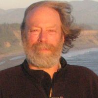 An older white man looking at the camera with gray beard and mustache, and receding hairline, wearing a black zip up jacket, standing on a cliff overlooking a rugged coastline,