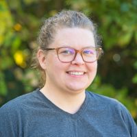 A white woman with short curly hair shaved on one side and a nosering, wearing a small gold necklace and black jacket, standing outside, looking at the camera and smiling